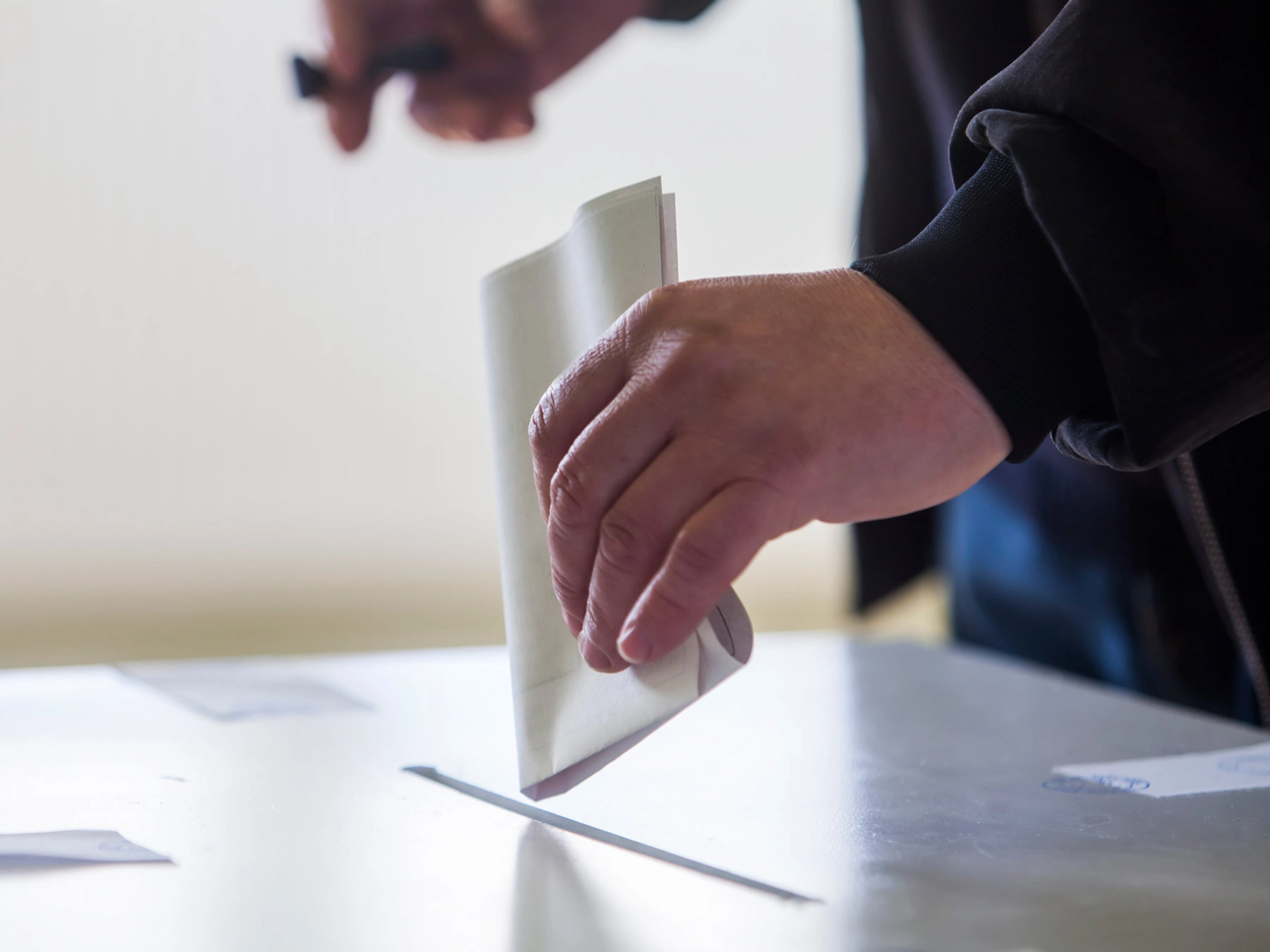 Jemand steckt mit der linken Hand einen gefalteten Wahlzettel in ein Wahlurne.
