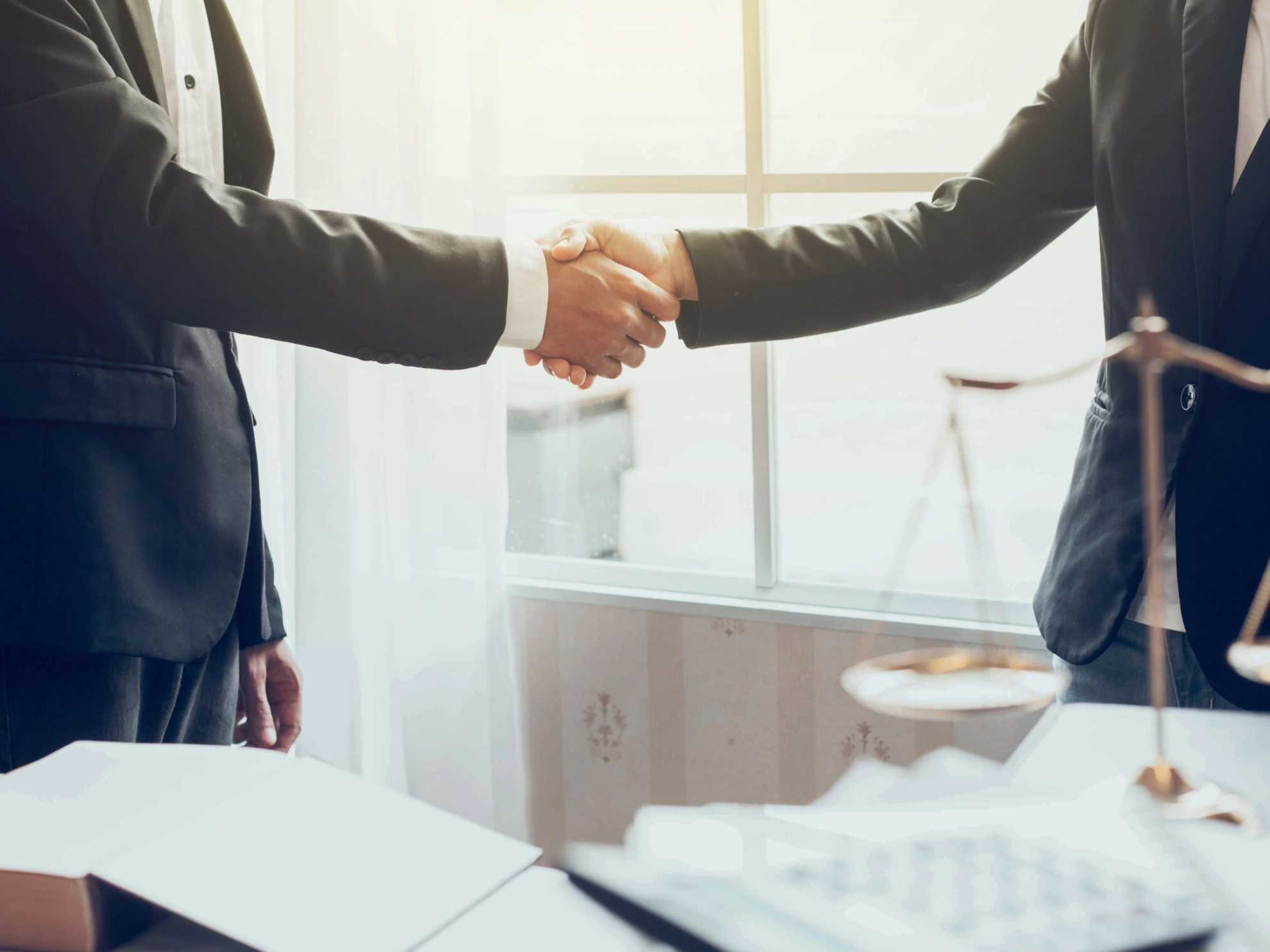 Zwei Männer schütteln sich die Hand neben einer Richterwaage auf dem Tisch, dort sind auch Akten zu sehen