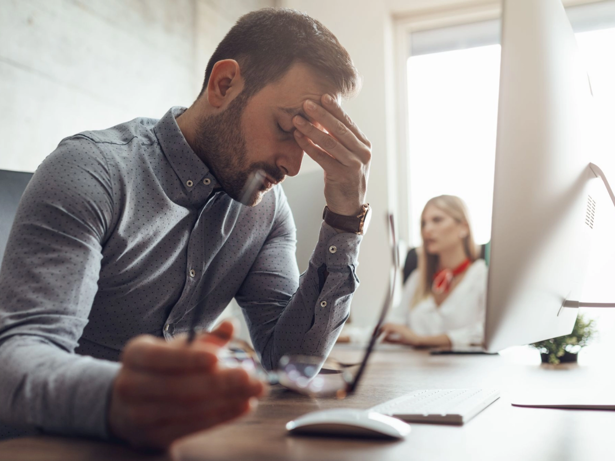 Junger Mann mit Brille reibt sich gestresst an seinem Arbeitsplatz die Augen